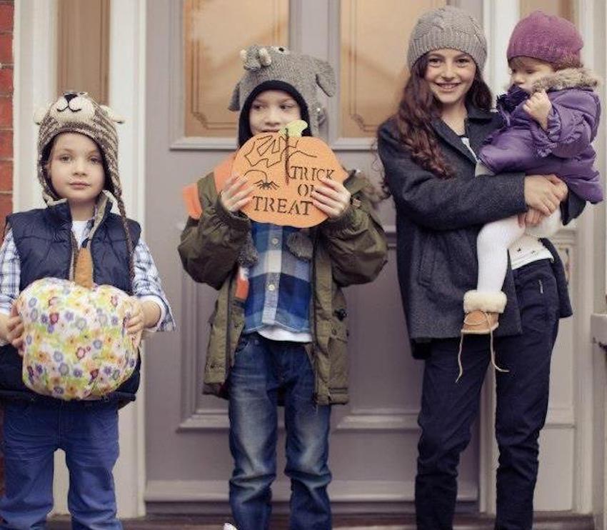 Four kids outside a front door dressed in winter clothes and hats holding Halloween props