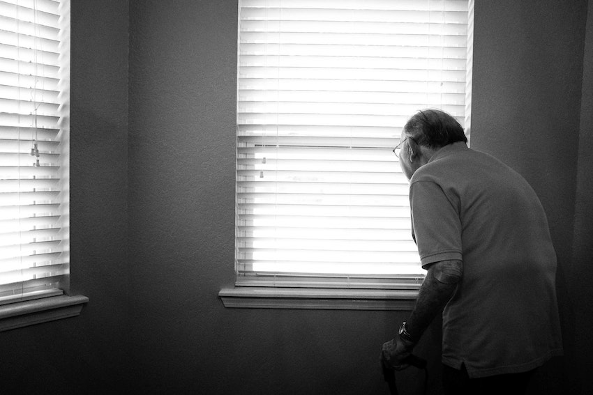 Elderly man standing at window with blind down