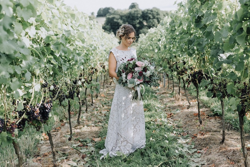 Bride holding bouquet standing between vines with dark grapes on