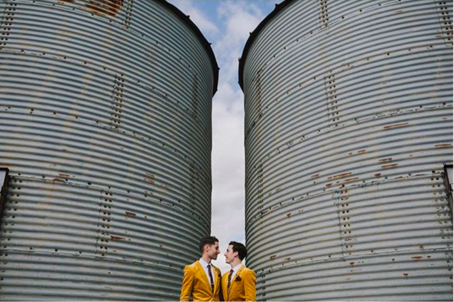 Image of gay couple in matching mustard suits standing by two huge water tanks looking in to each others eyes