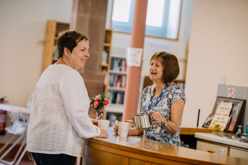 two women standing talking in a communal indoor space, one pouring tea for them both