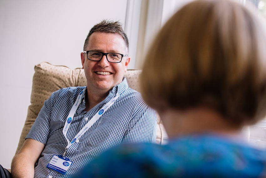 Cancer Care support worker - a white man with glasses and short greyish hair, talking to someone who is front of image but blurred